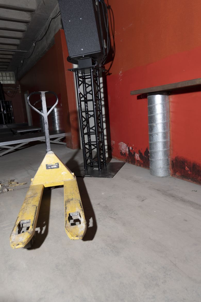 A large black PA speaker mounted on a truss against a red concrete wall.