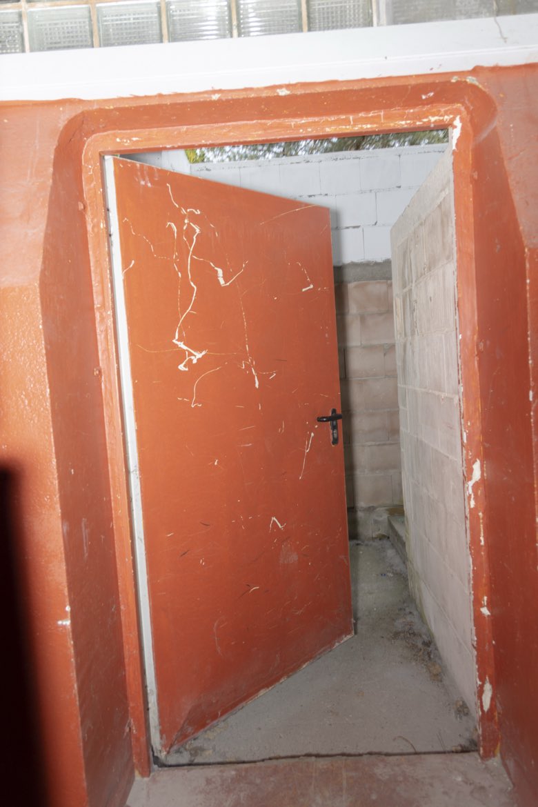 A weathered oxblood-red steel door opening onto a bare concrete stairwell.